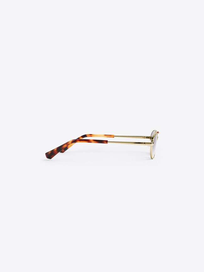 Oval eyeglasses with a tortoiseshell acetate frame and gold metal temples, shown from a side profile on a light gray background. 