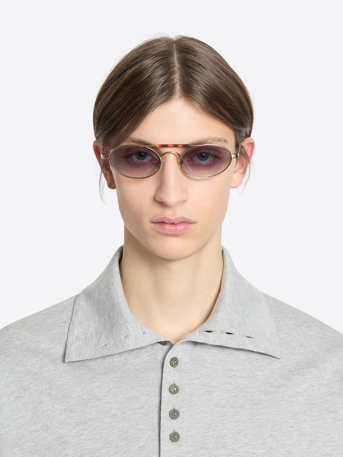 A model wearing tortoiseshell acetate sunglasses with oval tinted lenses and a light gray polo shirt, shot against a light gray background.