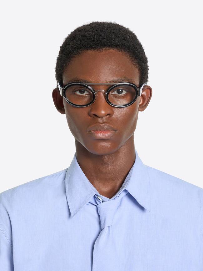 A model  wearing round black-rimmed eyeglasses with a distinctive double bridge and a light blue button-down shirt, shown against a light gray background.