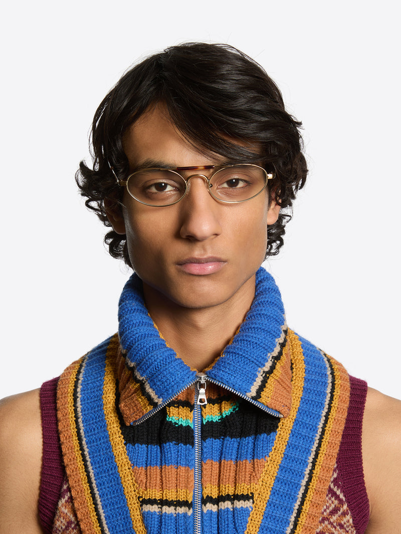 A man wearing oval metal-framed eyeglasses, a bright blue knit zip collar, and a colorful striped knit vest against a light gray background.