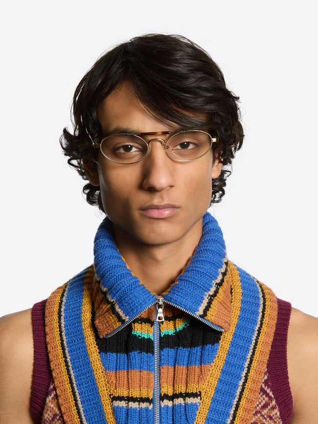 A man wearing oval metal-framed eyeglasses, a bright blue knit zip collar, and a colorful striped knit vest against a light gray background.