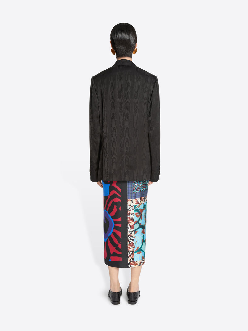 Back view of a person wearing a dark brown blazer and colorful patterned skirt on a white background