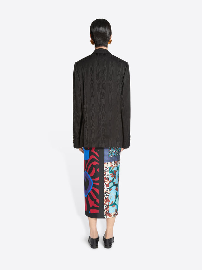 Back view of a person wearing a dark brown blazer and colorful patterned skirt on a white background