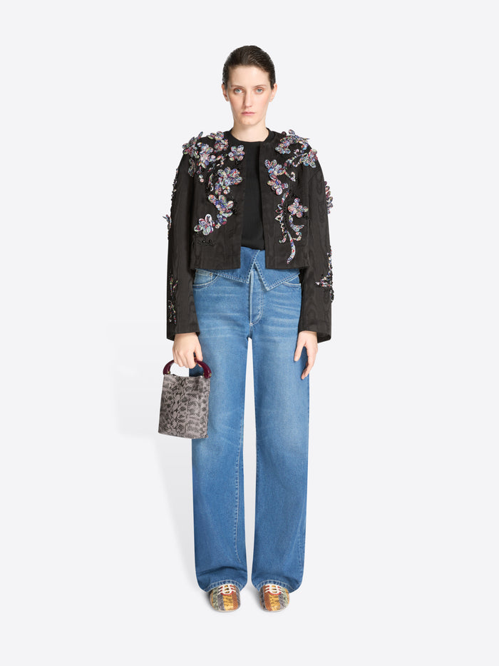 Woman wearing a black floral embroidered jacket and blue jeans, holding a handbag on a white background