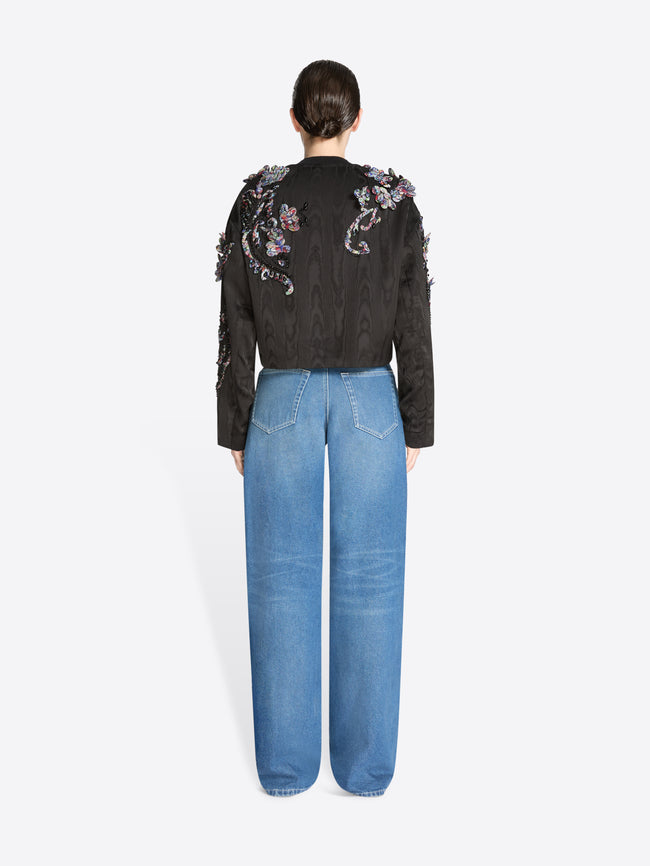 Back view of a person wearing a black floral embroidered jacket and blue jeans on a white background