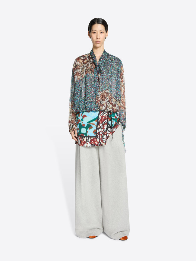 Model wearing a patterned blouse and light gray pants on a white background