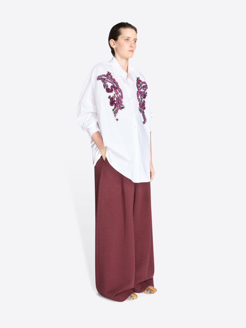 Woman wearing a white shirt with purple embroidery and burgundy pants on a white background