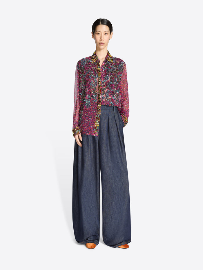 Model wearing a sheer patterned blouse and dark denim pants on a white background