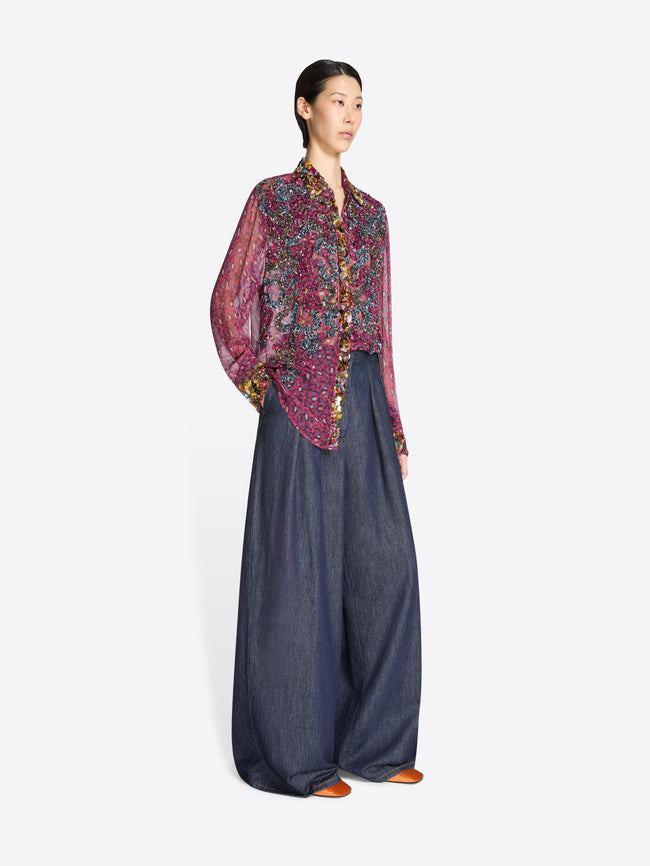 Woman wearing a sheer patterned blouse and dark denim pants on a white background