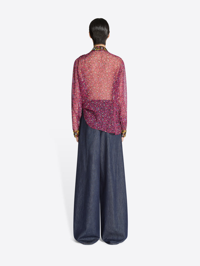 Back view of a person wearing a sheer pink top with a pattern and dark blue denim pants on a white background