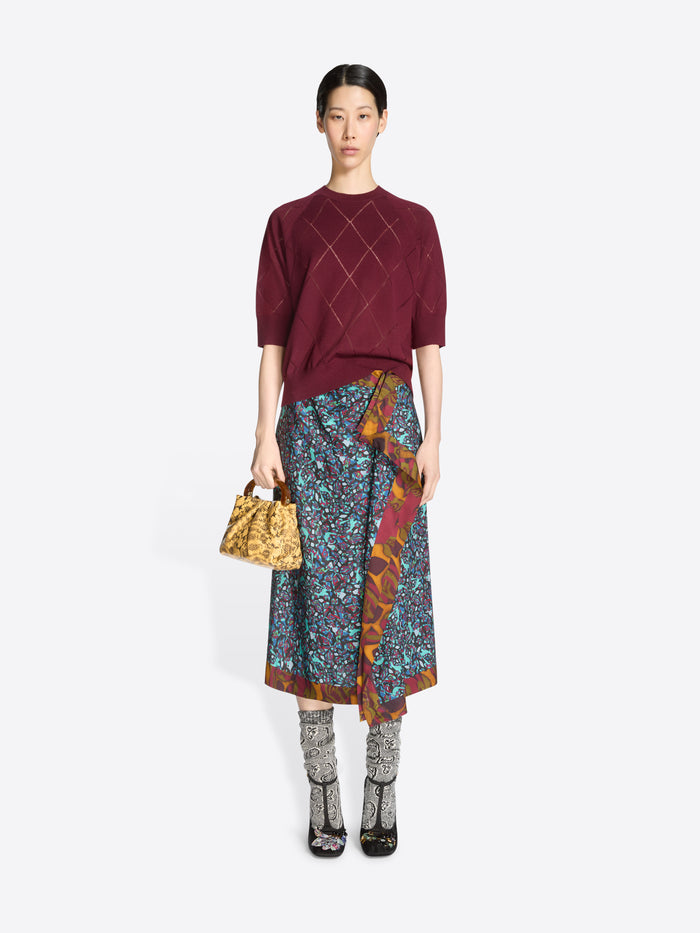 Woman wearing a burgundy knit top and patterned skirt with a yellow handbag on a white background