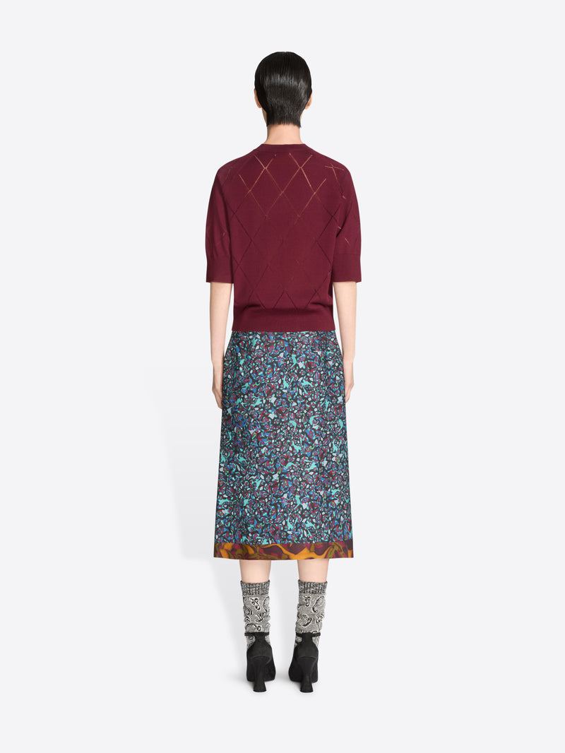 Back view of a person wearing a burgundy knit top and patterned skirt on a white background