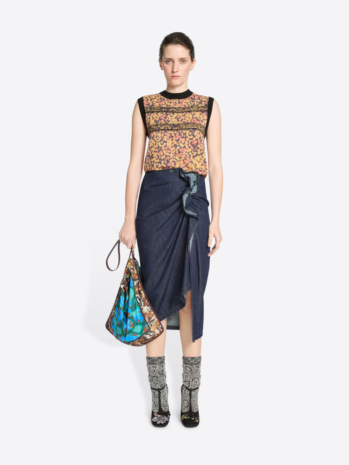 Woman wearing a patterned floral top and denim skirt holding a colorful bag on a white background