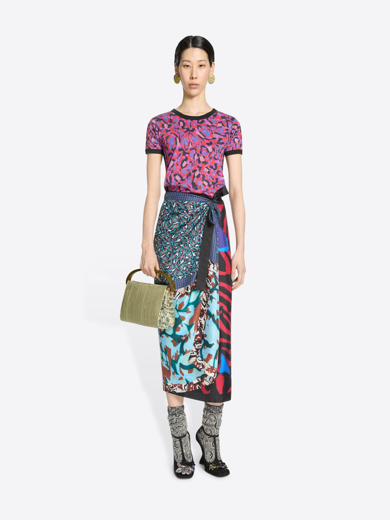 Model wearing a patterned top and skirt, holding a green handbag on a white background