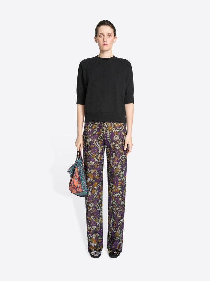 Woman wearing a black top and patterned pants holding a colorful bag on a white background
