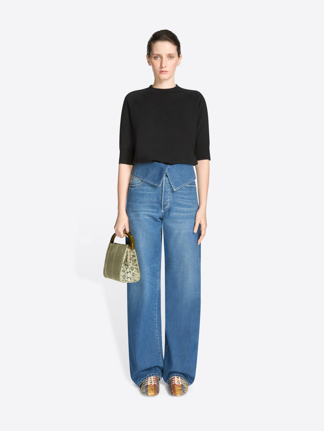 Woman wearing a black top and blue jeans holding a green handbag on a white background