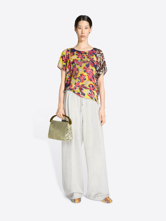 Woman wearing a colorful floral blouse and light-colored pants, holding a green handbag on a white background