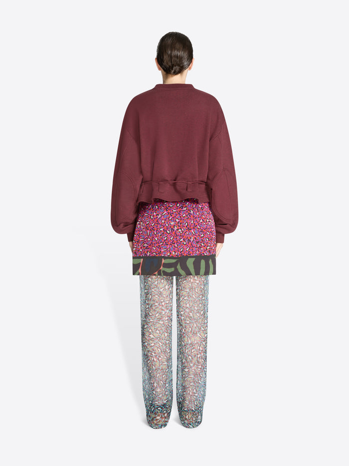 Back view of a person wearing a burgundy sweatshirt and patterned pants on a white background