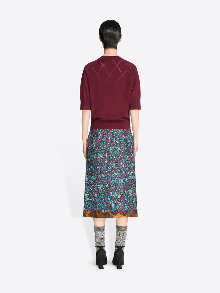Back view of a person wearing a burgundy knit top and patterned skirt on a white background