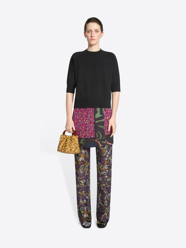 Woman wearing a black top, skirt and colorful pants holding a patterned handbag on a white background