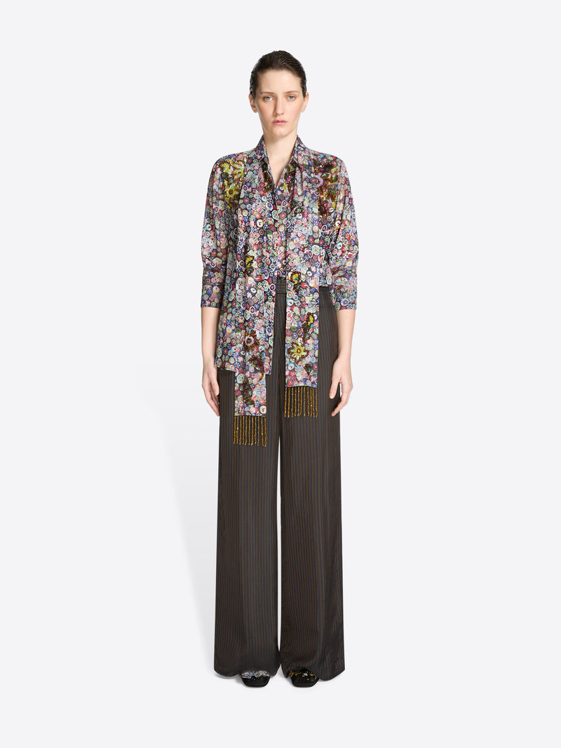 Woman wearing a floral blouse with a multicolored matching scarf with gold accents and wide-leg pants on a white background