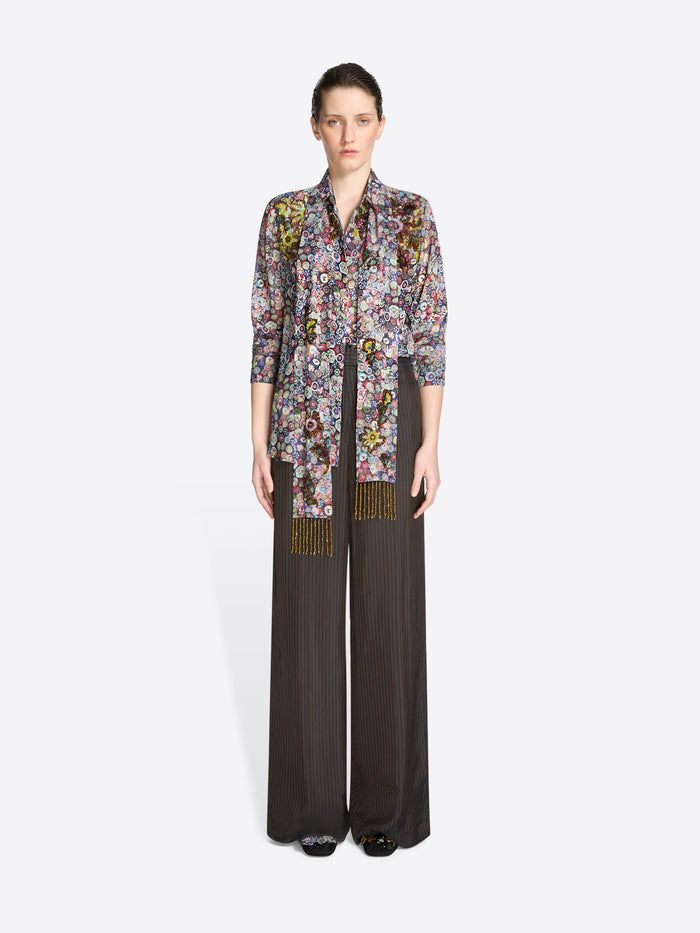 Woman wearing a floral blouse with a multicolored matching scarf with gold accents and wide-leg pants on a white background