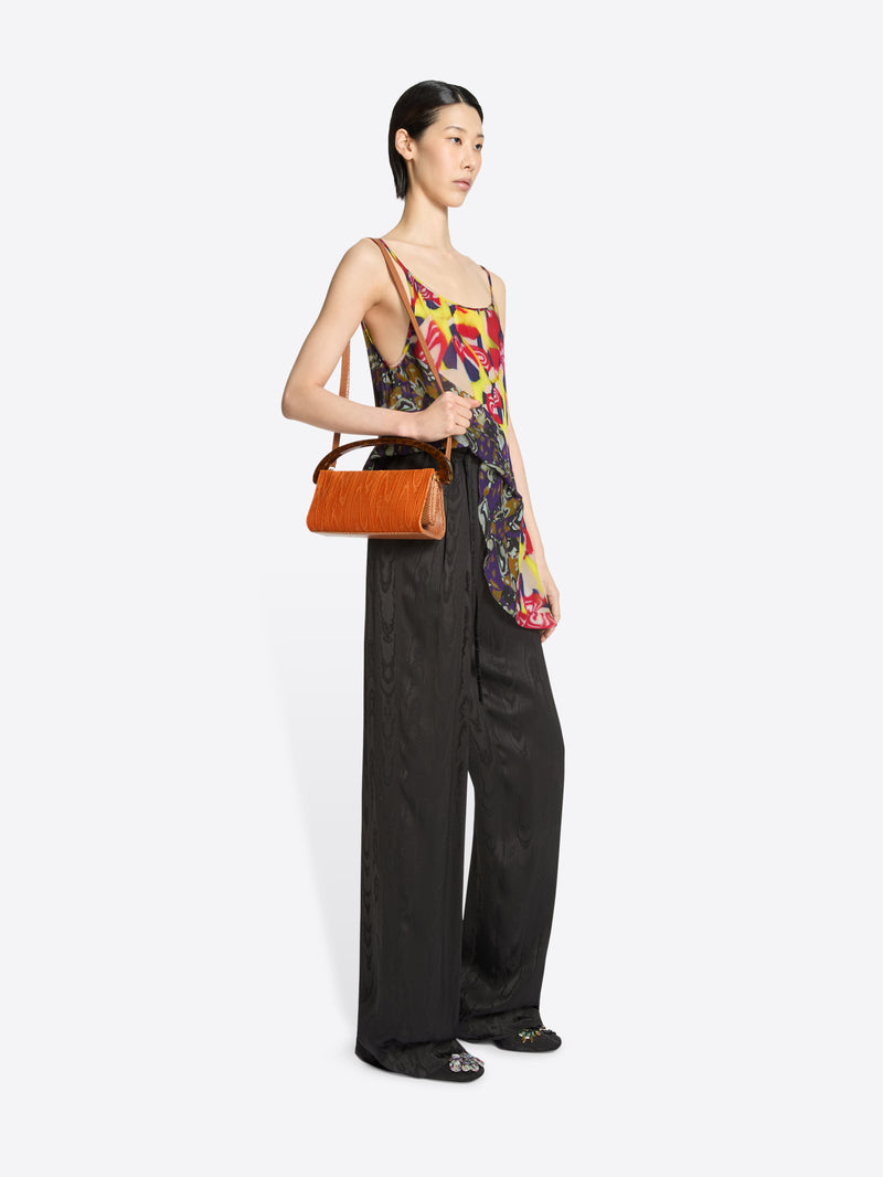Woman holding an orange handbag wearing a colorful top and dark brown pants on a white background