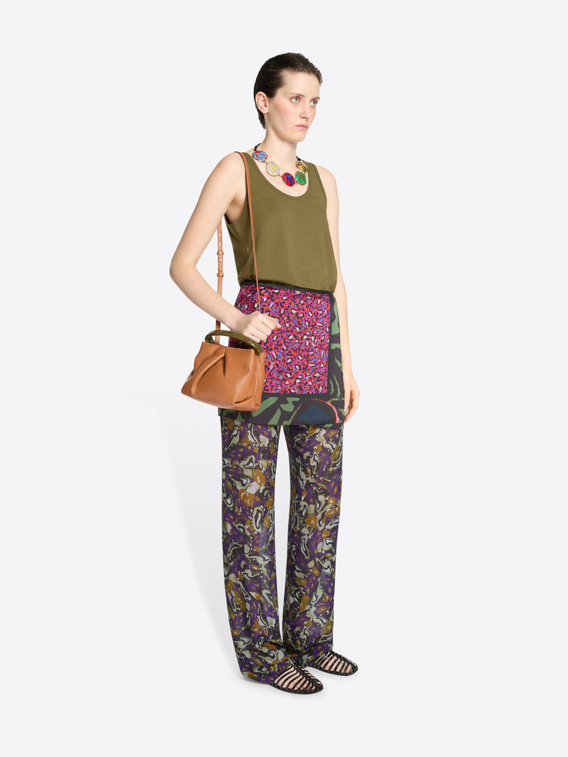 Woman wearing a green sleeveless top, skirt and patterned pants, holding an orange handbag on a white background