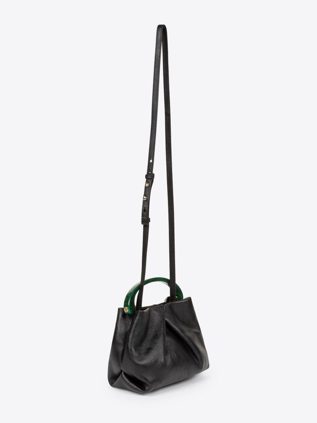 A black leather shoulder bag with a long strap, structured slouchy body, and contrasting dark green circular top handle, shown against a light gray background.