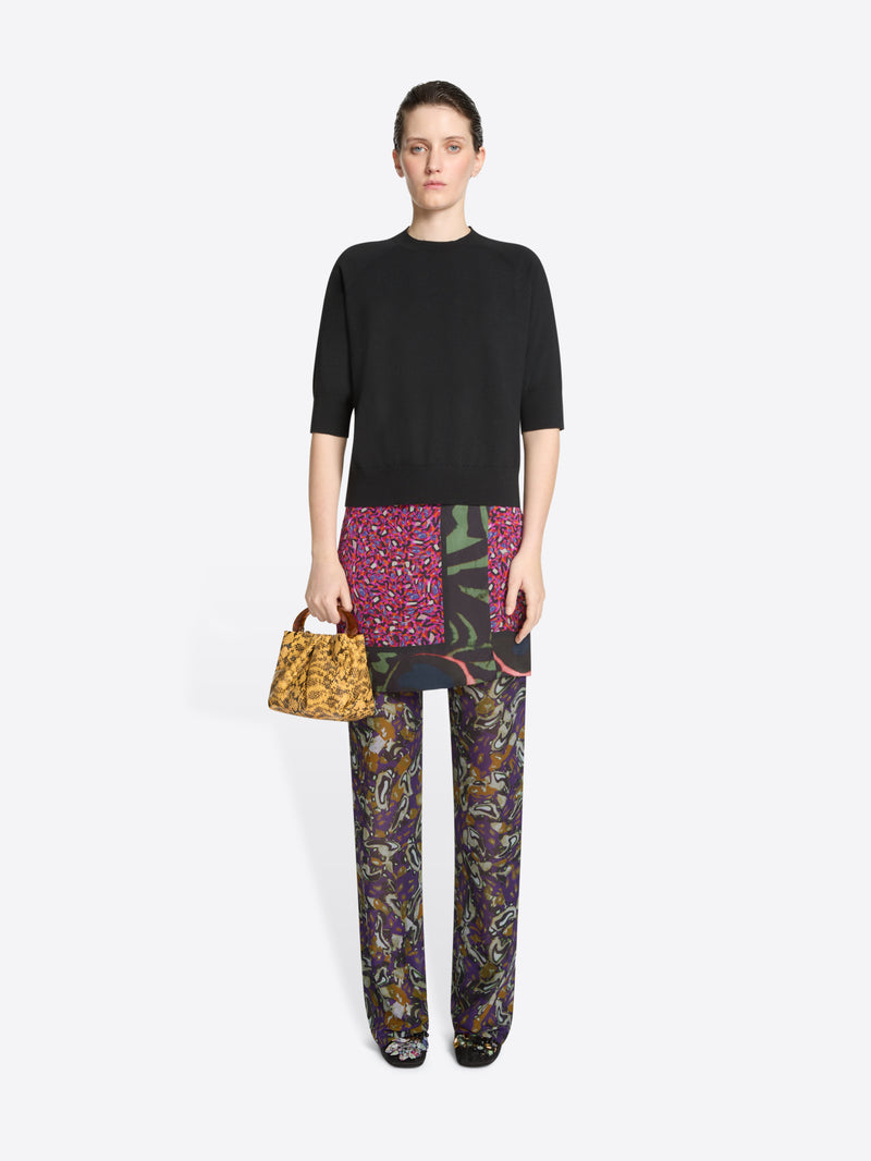 Woman wearing a black top, skirt and colorful pants holding a patterned handbag on a white background