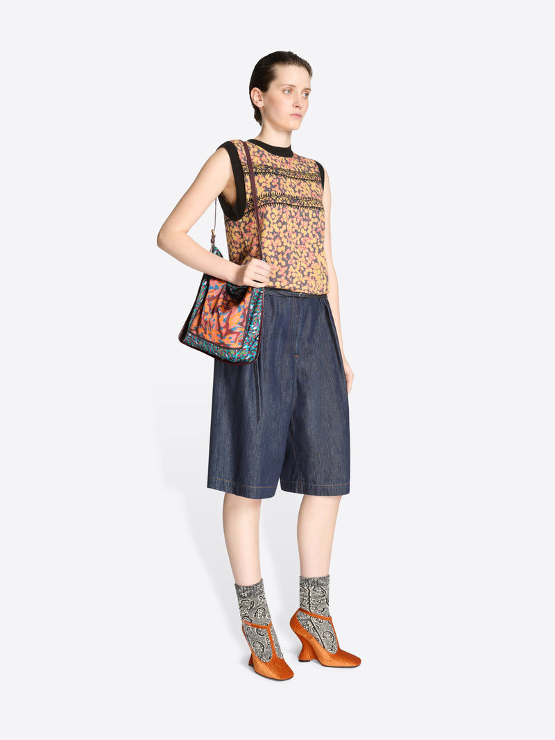 Woman wearing a sleeveless patterned top and denim shorts with a colorful bag on a white background