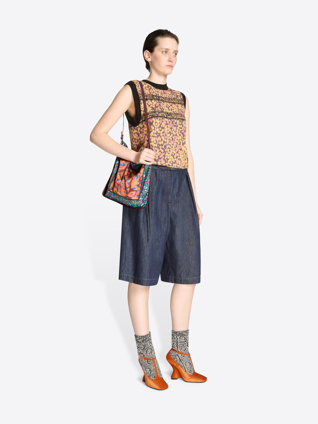 Woman wearing a sleeveless patterned top and denim shorts with a colorful bag on a white background