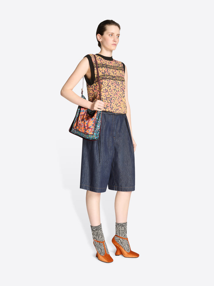 Woman wearing a sleeveless patterned top and denim shorts with a colorful bag on a white background