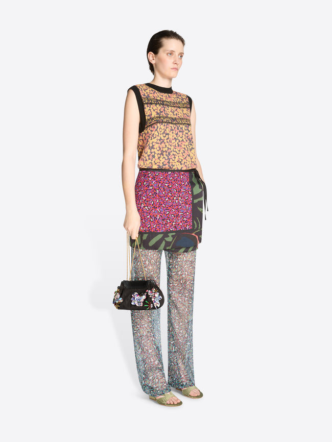 Model wearing a patterned outfit with a skirt, top and pants holding a black handbag on a white background