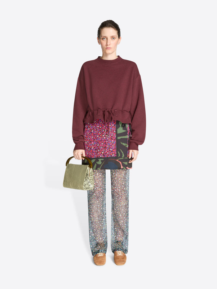 Person wearing a burgundy sweatshirt and patterned pants holding a green bag on a white background