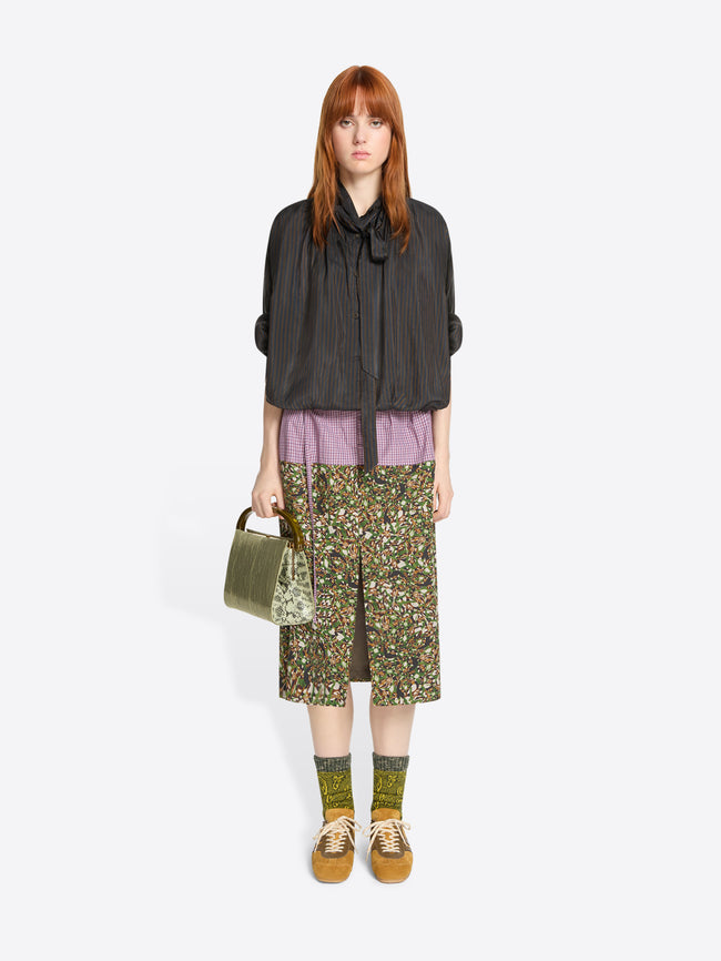 Woman wearing a brown shirt, patterned skirt, and yellow mustard shoes holding a green bag on a white background