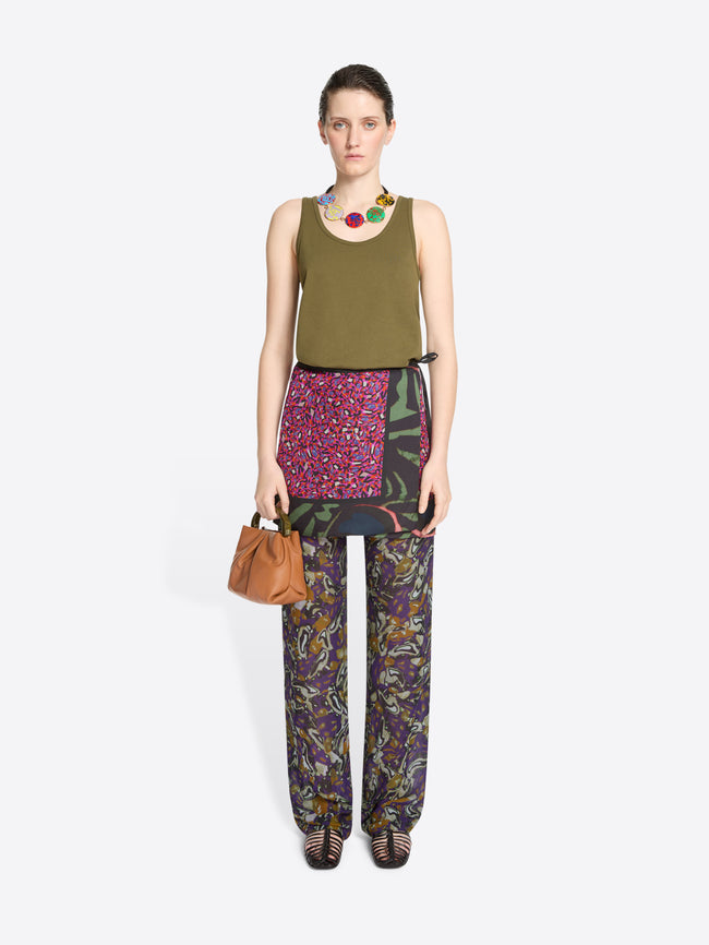 Woman wearing a green top, skirt patterned pants and black strappy sandals with a matching bag on a white background