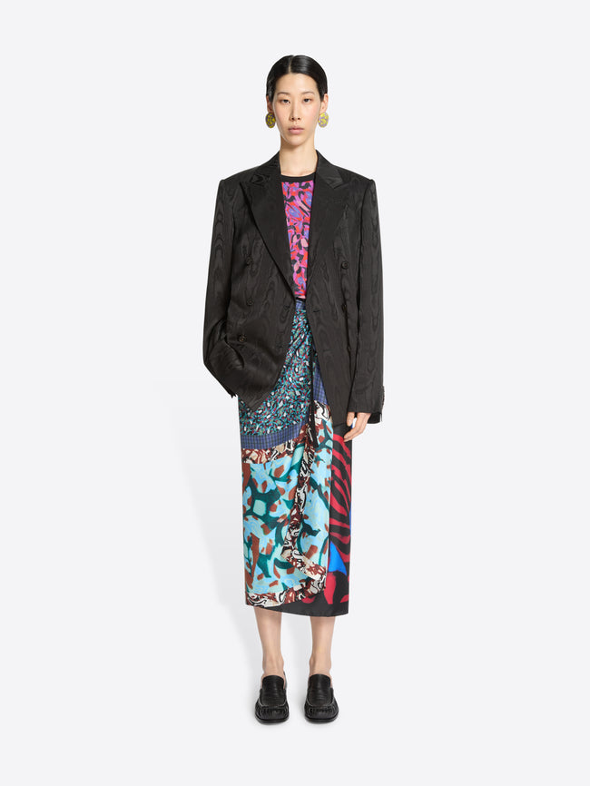 Woman wearing a dark brown blazer over a colorful top and skirt with black loafers on a white background