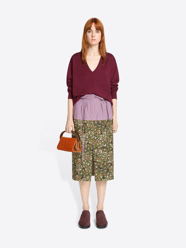 Woman wearing a burgundy sweater, purple shirt, and floral skirt with a orange handbag on a light grey background