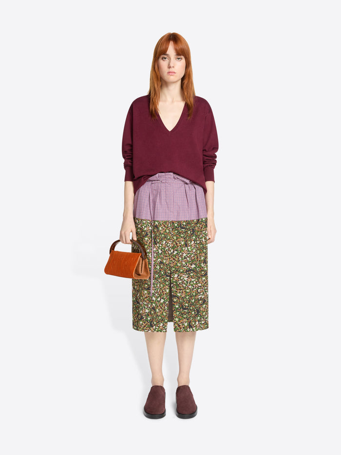 Woman wearing a burgundy sweater, purple shirt, and floral skirt with a orange handbag on a light grey background