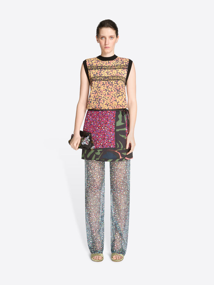 Model wearing a colorful outfit with a patterned top, skirt, sheer pants and green mules on a white background