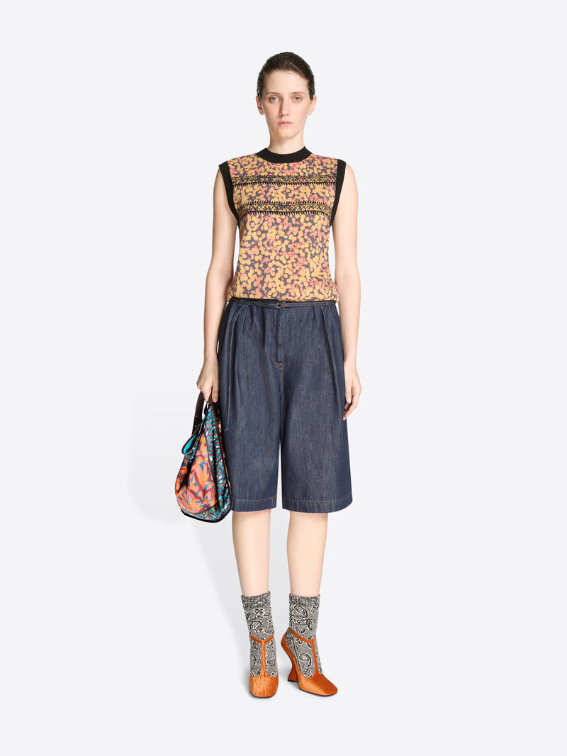 Woman wearing a sleeveless floral top, denim shorts and orange high-heeled shoes on a white background