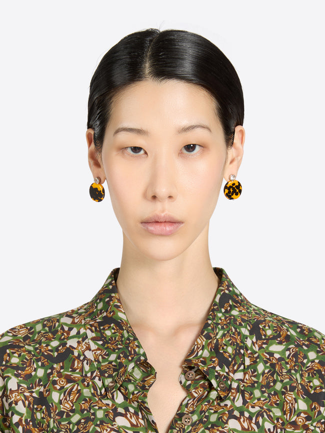 Woman wearing round earrings and a patterned button-up blouse with a plain background