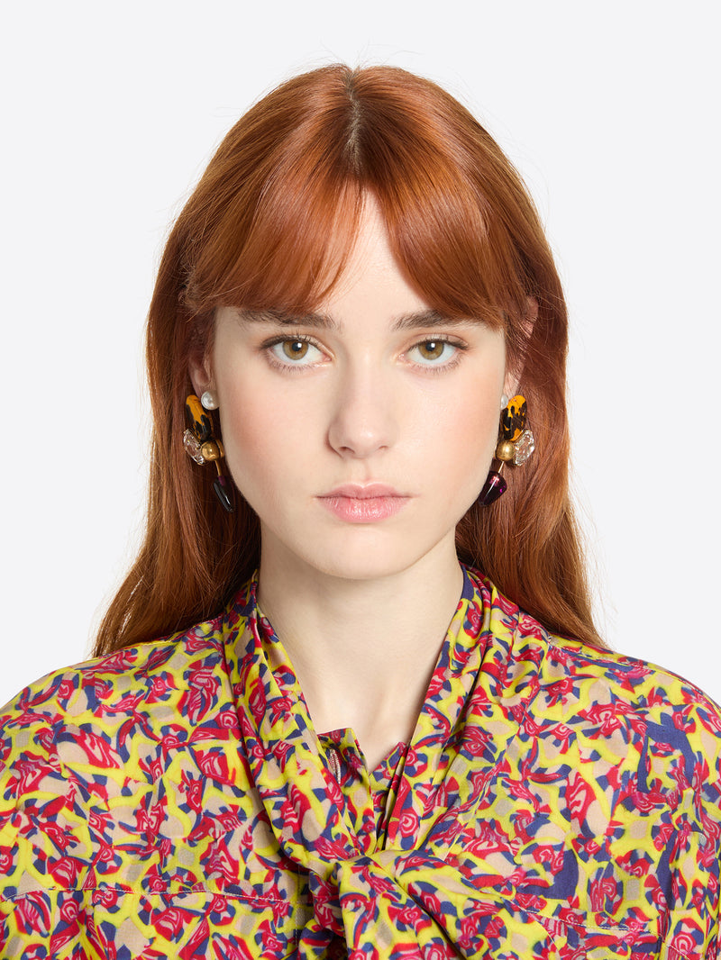 Woman wearing a pair of earrings with colorful gemstones and a colorful floral blouse against a white background