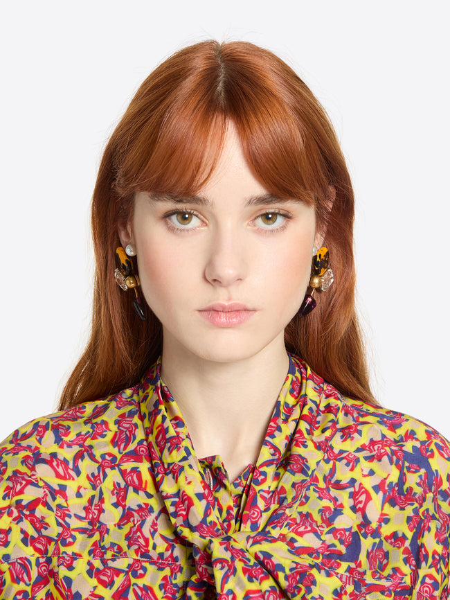 Woman wearing a pair of earrings with colorful gemstones and a colorful floral blouse against a white background