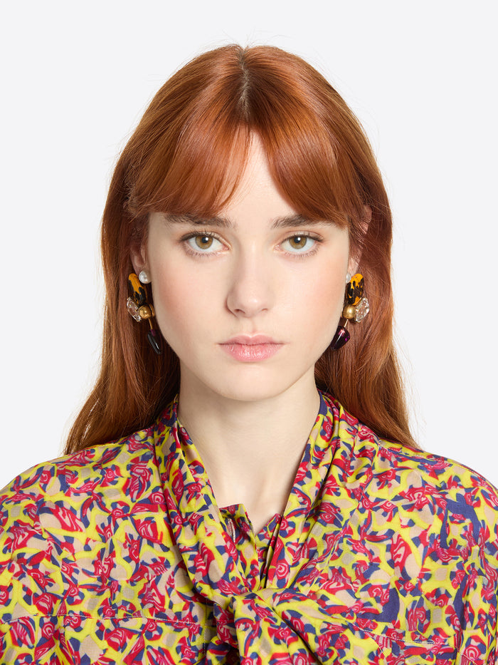 Woman wearing a pair of earrings with colorful gemstones and a colorful floral blouse against a white background