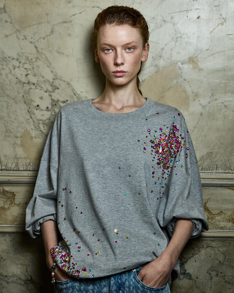 Woman wearing a gray sweater with colorful sequin embellishments against a textured wall.