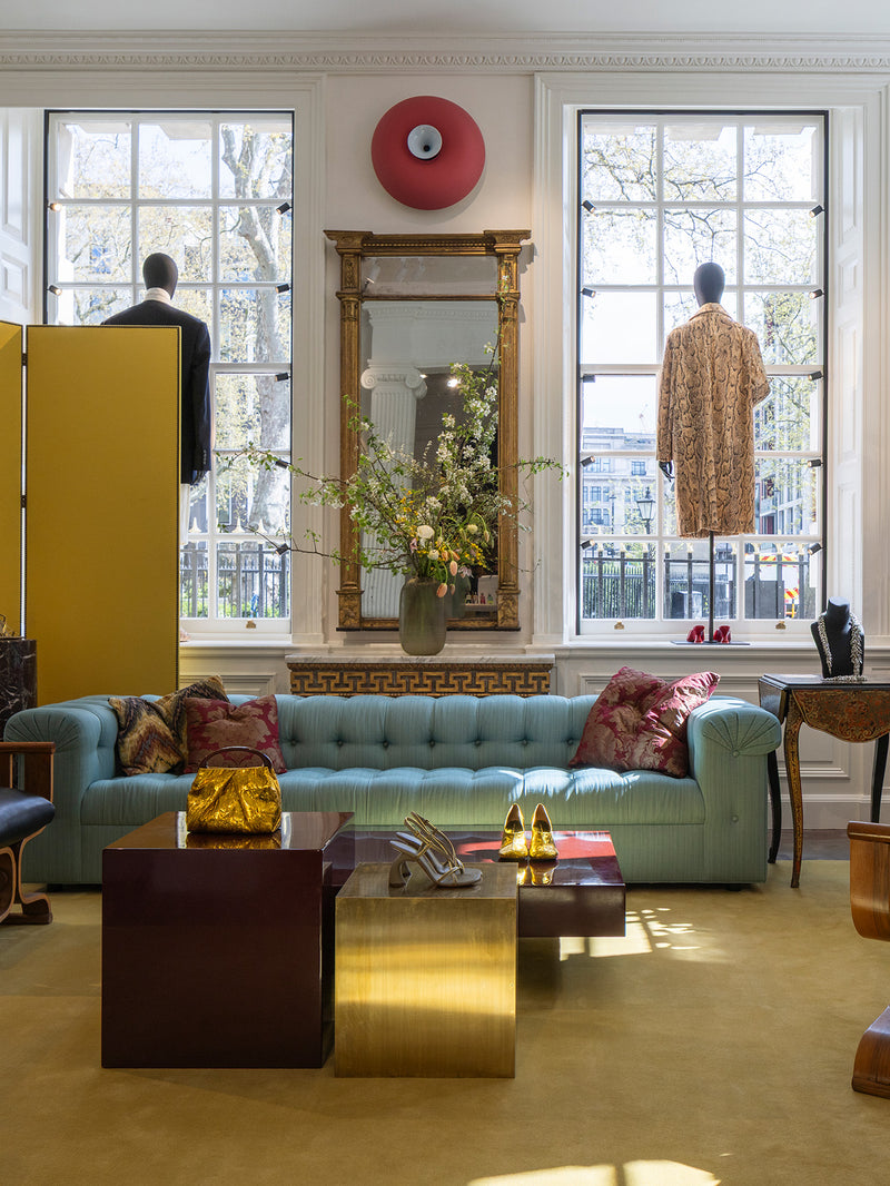 A styled interior space with a light blue tufted sofa, patterned pillows, a gold table, large windows, and mannequins wearing garments. 