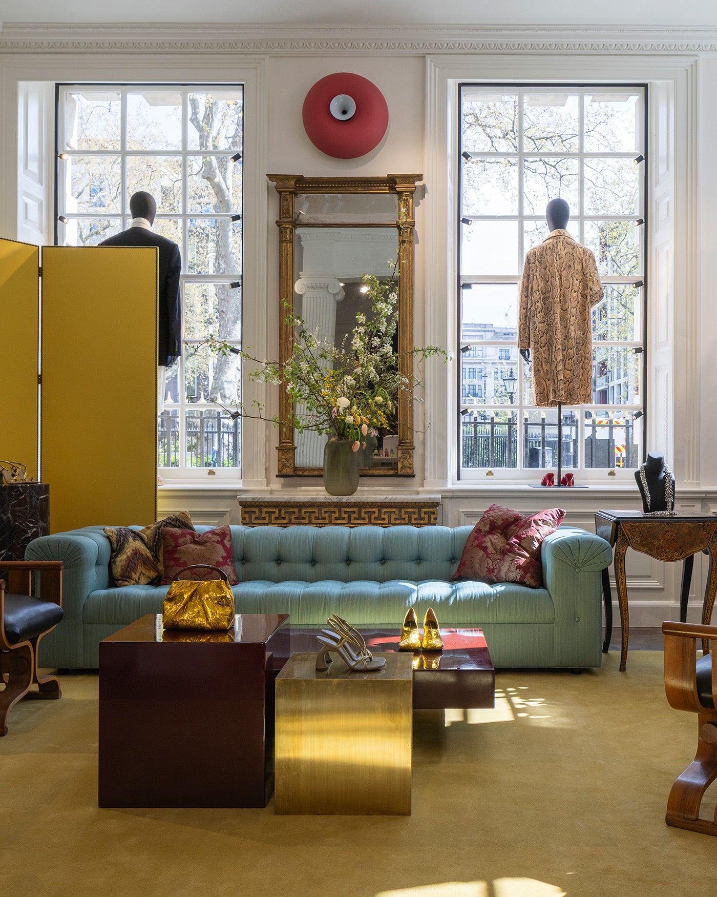 A styled interior space with a light blue tufted sofa, patterned pillows, a gold table, large windows, and mannequins wearing garments. 
