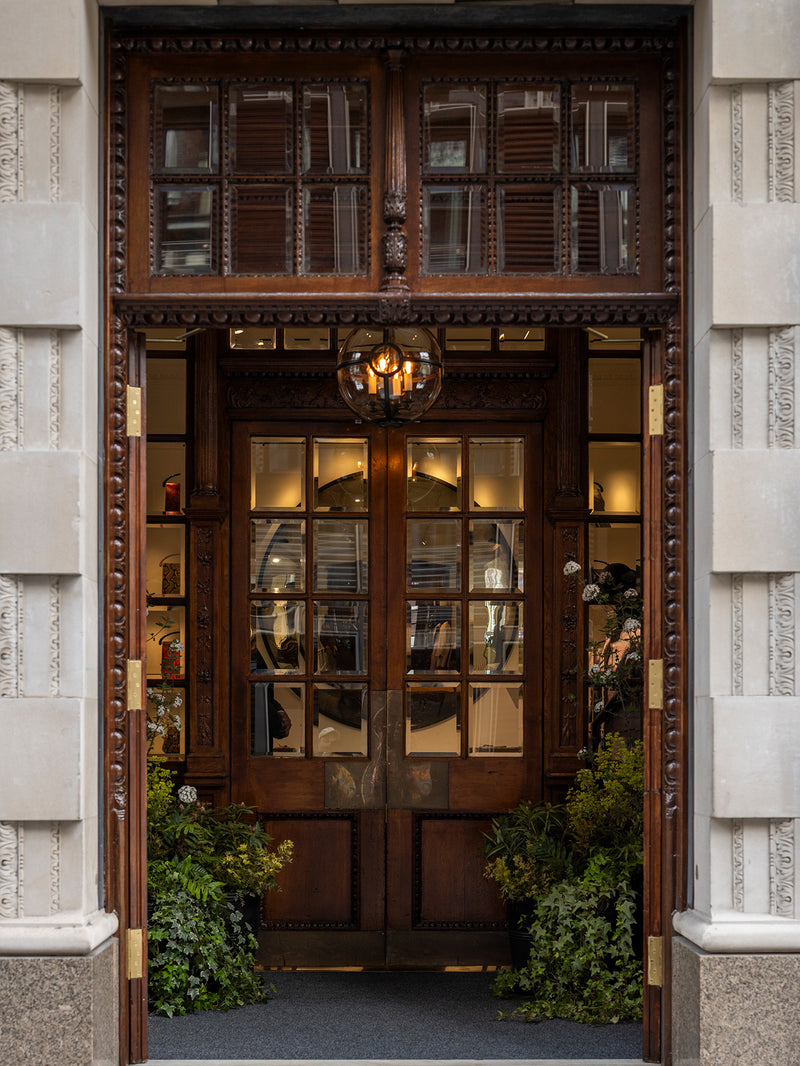 An ornate dark wood double doorway with decorative carved trim, glass panes, brass hinges, and a hanging lantern light, set in a light stone building facade with greenery at the base.